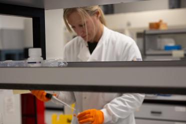 Scientist wearing orange safety gloves pipettes liquid into the top left corner of a well-plate