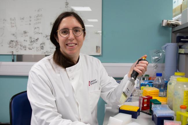 Dr Sarah Guiziou photographed siting down facing the camera, in a white lab coat holding a multi channel pipette. 